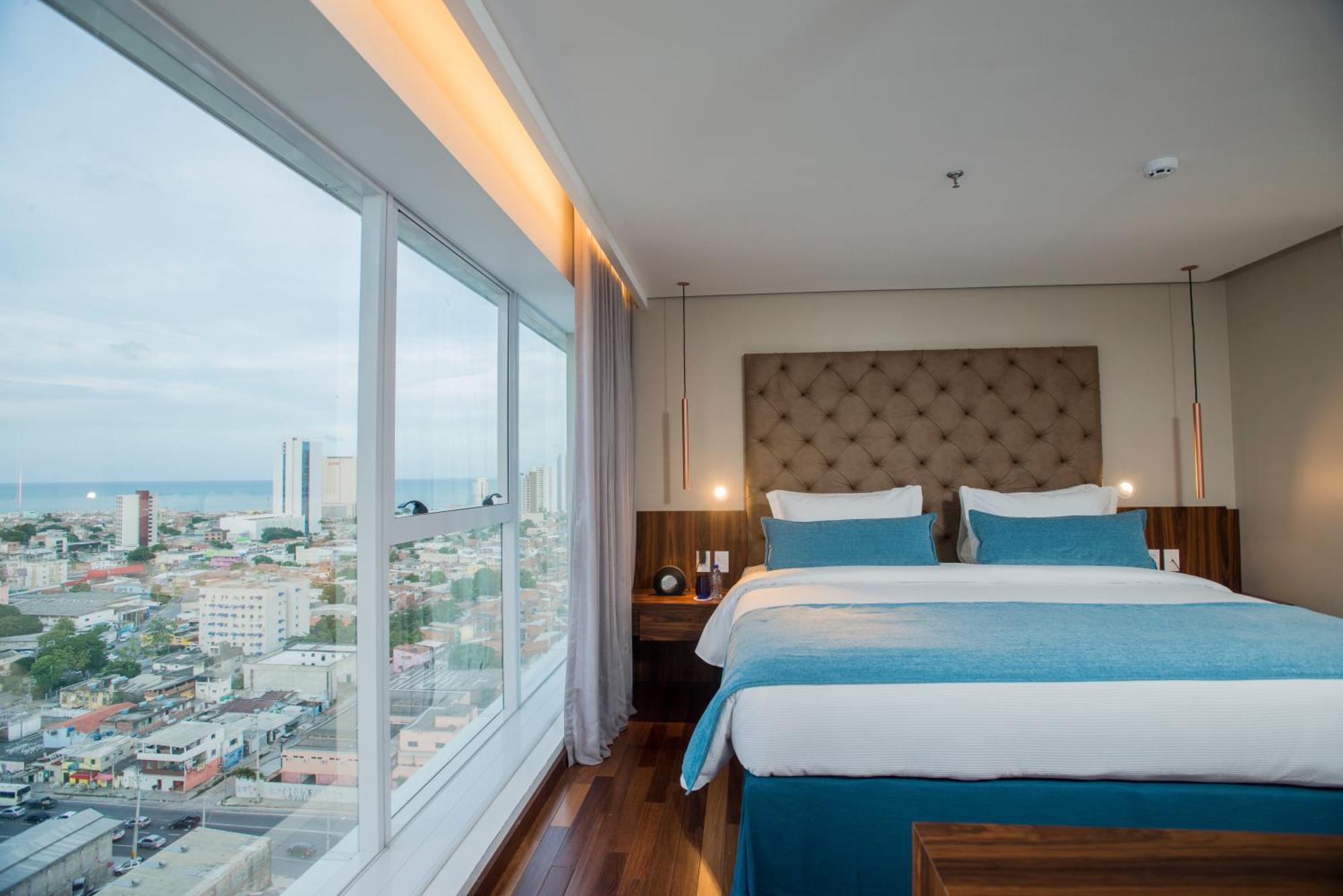 Hotel Luzeiros 헤시피 외부 사진 The photo shows a modern hotel room featuring a large window with a city view. Inside, there are two neatly made beds with light blue and white bedding, flanked by wooden nightstands. The room has a contemporary design, with a padded headboard and so